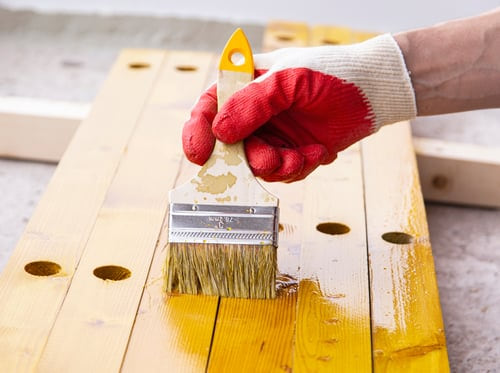 Gloved hand applying primer with a wide brush to bare wood boards during surface preparation